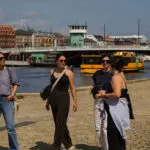 Four women stroll along a sunny waterfront promenade in Christianshavn, with the neighborhood's highlights—a bridge, buildings, and yellow water taxis—creating a picturesque backdrop filled with hidden gems.