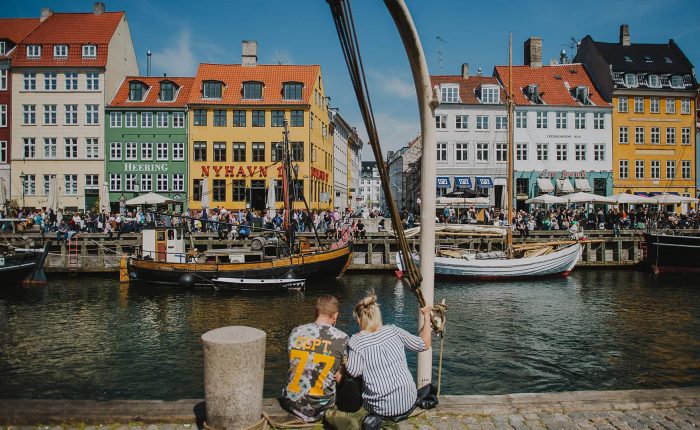Two people sit by a canal in Nyhavn, Copenhagen, with colorful historic buildings and boats in the background on a sunny day.
