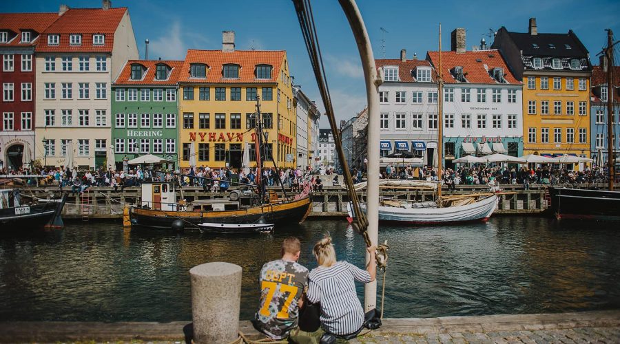 Two people sit by a canal in Nyhavn, Copenhagen, with colorful historic buildings and boats in the background on a sunny day.